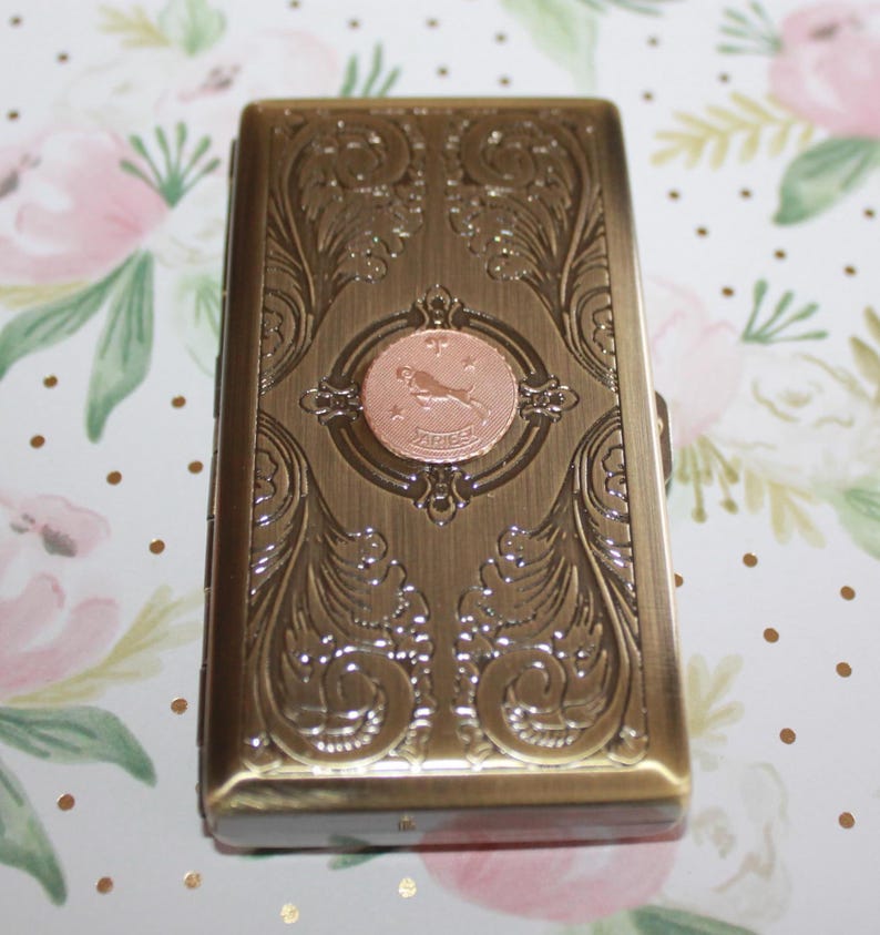 May include: A rectangular, antique brass-colored case with ornate floral and scrollwork designs. A circular copper-toned emblem in the center features a leaping figure and the word "ARIES". The case has a hinged lid and a clasp.