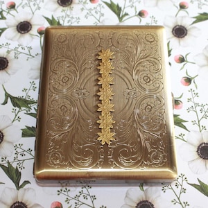 May include: A rectangular brass-toned metal cigarette case featuring an intricate embossed floral scrollwork pattern across the surface, accented by a vertical row of six small, raised, connected flower-shaped metal embellishments.
