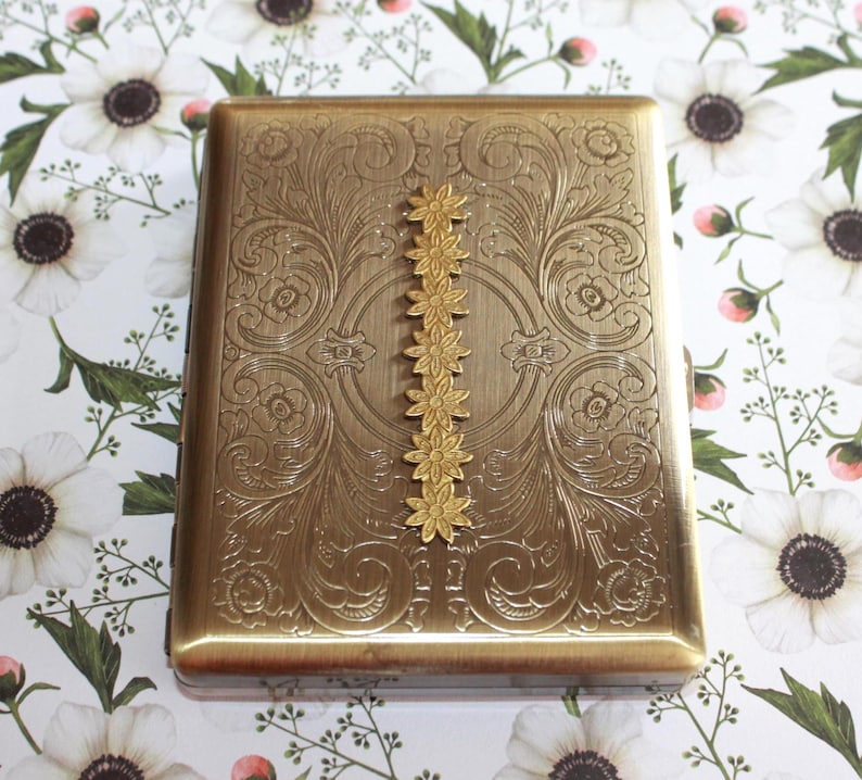 May include: A rectangular brass-toned metal cigarette case featuring an intricate embossed floral scrollwork pattern across the surface, accented by a vertical row of six small, raised, connected flower-shaped metal embellishments.