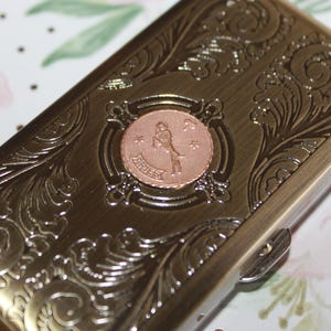 May include: A rectangular, bronze-colored metal case with ornate, swirling floral engravings. The center features a circular, copper-toned emblem with the word "ARIES" and a figure. The case has a clasp closure.