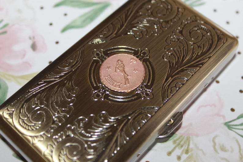 May include: A rectangular, bronze-colored metal case with ornate, swirling floral engravings. The center features a circular, copper-toned emblem with the word "ARIES" and a figure. The case has a clasp closure.