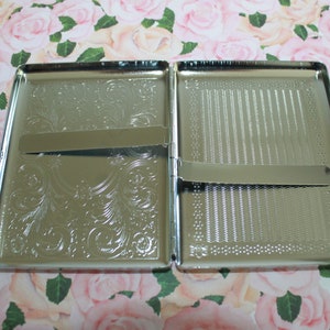 May include: An open, silver cigarette case. One side features an engraved floral pattern, the other a textured design. The rectangular case has a hinged opening and a metal clasp to secure cigarettes. The case is made of metal.