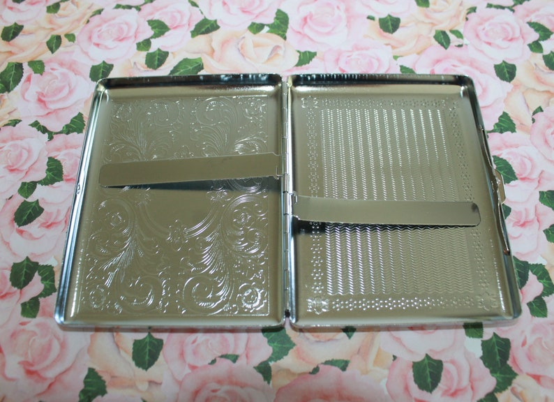 May include: An open, silver cigarette case. One side features an engraved floral pattern, the other a textured design. The rectangular case has a hinged opening and a metal clasp to secure cigarettes. The case is made of metal.