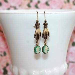 May include: A pair of drop earrings featuring a metallic bronze-toned hand motif suspended from a hook, with a small oval-shaped green and white cameo pendant dangling from the wrist of each hand, showcasing a profile portrait of a person.