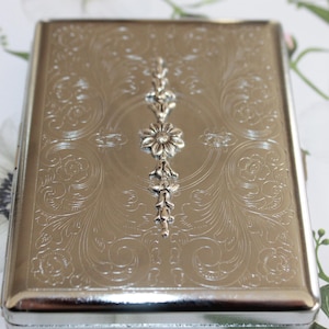 May include: Silver cigarette case with an ornate floral design and a silver flower accent.