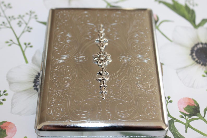 May include: Silver cigarette case with an ornate floral design and a silver flower accent.