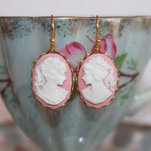 May include: A pair of gold earrings with pink cameo pendants featuring white porcelain profile portraits of women.