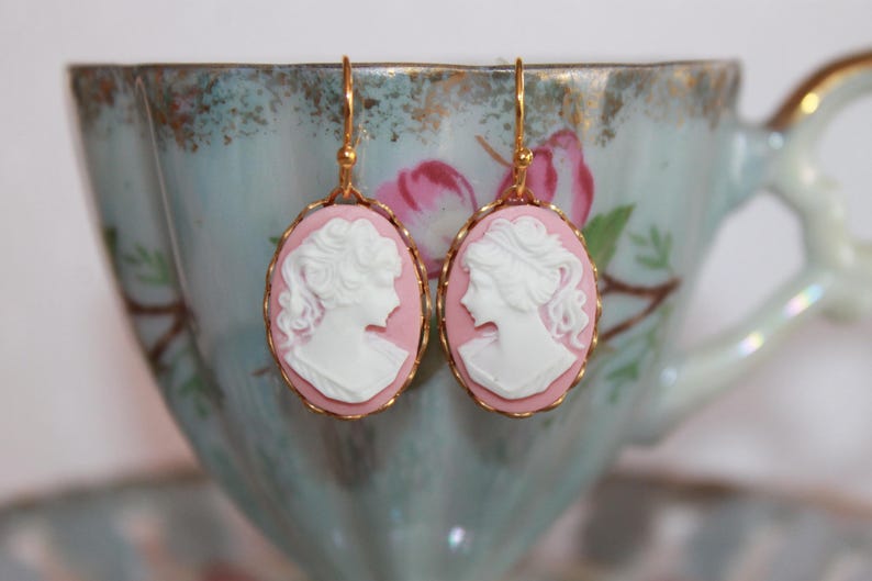 May include: A pair of gold earrings with pink cameo pendants featuring white porcelain profile portraits of women.
