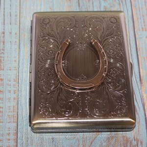 May include: A rectangular, metallic cigarette case with an embossed floral design and a horseshoe accent. The case is a muted bronze color and sits on a light blue and brown wooden surface.