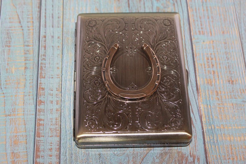 May include: A rectangular, metallic cigarette case with an embossed floral design and a horseshoe accent. The case is a muted bronze color and sits on a light blue and brown wooden surface.