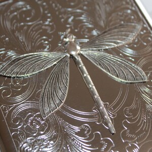 May include: Silver-colored metal case with an embossed dragonfly design. The case has a detailed floral pattern and a hinged closure. The dragonfly is the focal point, adding a decorative element to the case.