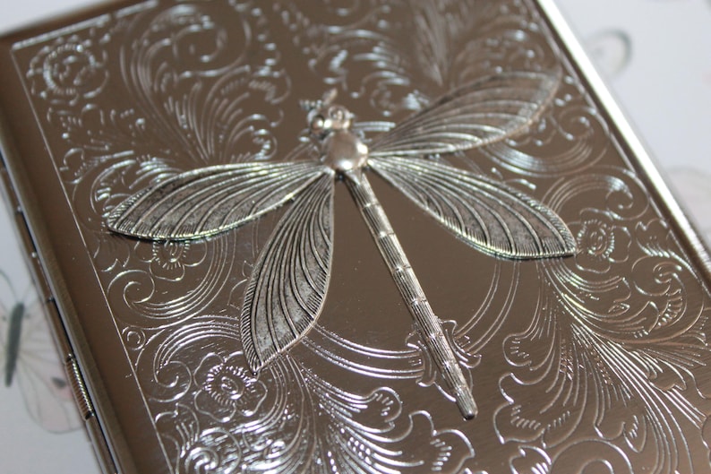 May include: Silver-colored metal case with an embossed dragonfly design. The case has a detailed floral pattern and a hinged closure. The dragonfly is the focal point, adding a decorative element to the case.