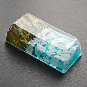May include: A rectangular, clear resin keycap with a blue and white wave design and a green and brown island design.