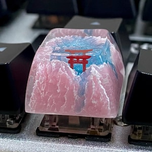 May include: A custom keycap featuring a Japanese torii gate design. The keycap has a pink mountain range and blue sky background. The torii gate is red. The keycap is on a mechanical keyboard.