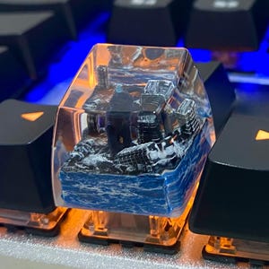 Puede incluir: Una tecla de teclado personalizada con una escena oceánica en miniatura. La tecla transparente tiene una base oceánica azul y blanca, una pequeña figura y edificios. La tecla se ilumina desde abajo con un brillo naranja, sobre un teclado negro.