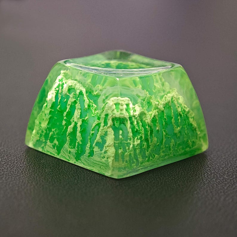 May include: A square, translucent green keycap with a mountain range design. The keycap is made of resin and features a detailed, textured landscape in shades of green and white. The keycap is designed for a mechanical keyboard.