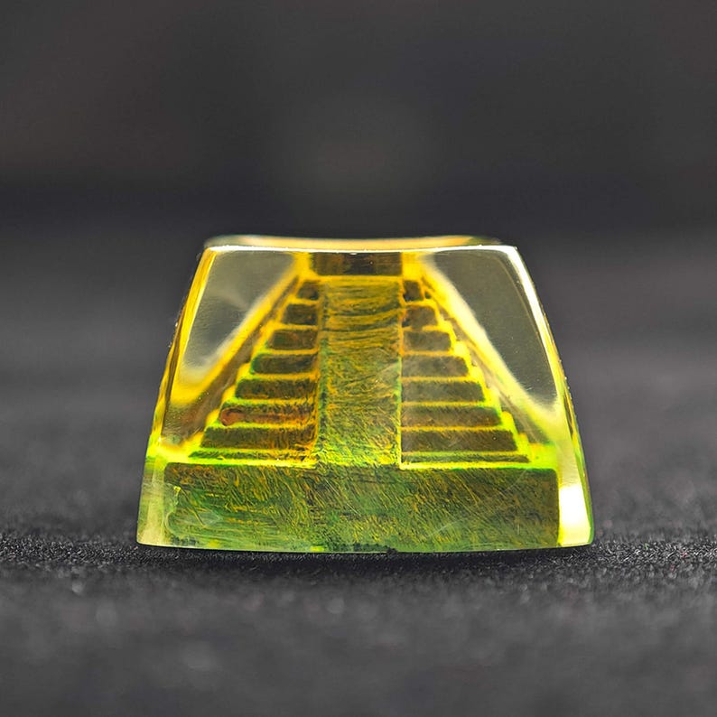 May include: A transparent, rectangular keycap with a yellow and green pyramid design. The keycap has a stepped pyramid design with a yellow and green gradient. The keycap is made of a clear resin material.