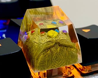 May include: A clear resin keycap with a gold-colored Infinity Gauntlet design. The Gauntlet has six colorful gems.