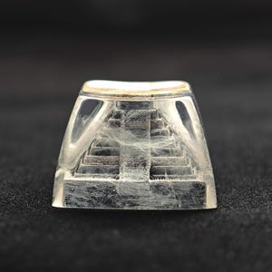 May include: A clear, rectangular keycap with a miniature pyramid design inside. The pyramid is gray and has visible steps. The keycap has a gold-colored top edge and is set against a dark background.