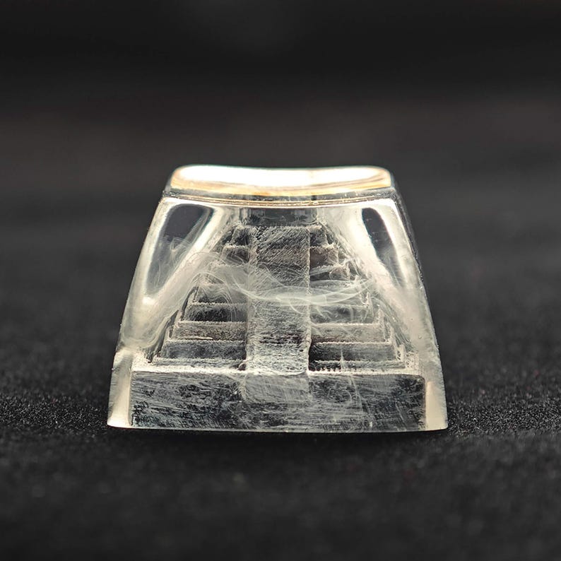 May include: A clear, rectangular keycap with a miniature pyramid design inside. The pyramid is gray and has visible steps. The keycap has a gold-colored top edge and is set against a dark background.