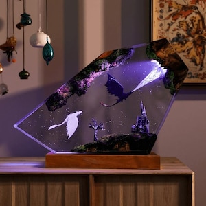 May include: A fantasy resin art piece depicting a dragon breathing fire, a white dragon silhouette, and a castle. The artwork is encased in a clear, angled resin block, mounted on a wooden base, and illuminated with purple light.
