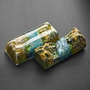May include: Two resin keycaps with a landscape design. The keycaps are a light blue, green, and brown color with a textured surface. The keycaps are made to look like a mountain range with a river running through it.