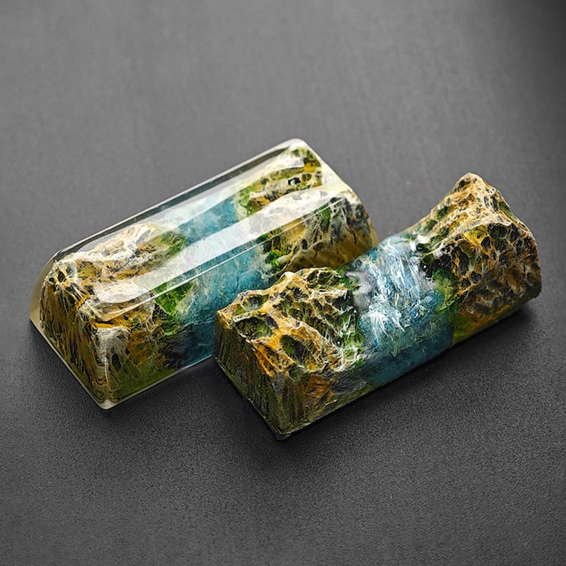 May include: Two resin keycaps with a landscape design. The keycaps are a light blue, green, and brown color with a textured surface. The keycaps are made to look like a mountain range with a river running through it.
