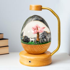 Könnte beinhalten: Ein Holzsockel mit einer durchsichtigen, eiförmigen Glaskuppel, die eine Miniatur-Waldszene mit einem rosa Baum, einem weißen Reh und Steinen enthält. Der Sockel hat einen goldenen Metallarm, der als Leuchte dient.