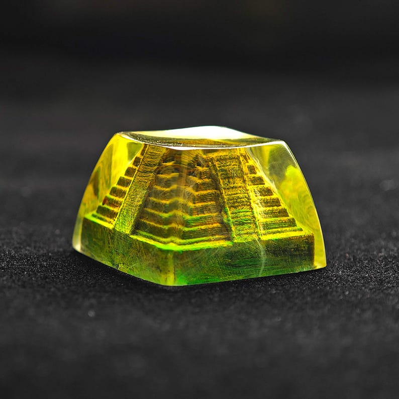 May include: A translucent, yellow-green resin keycap featuring a detailed, miniature pyramid structure. The pyramid has visible steps and is encased within the clear resin, creating a unique, artistic effect. The keycap is square-shaped.