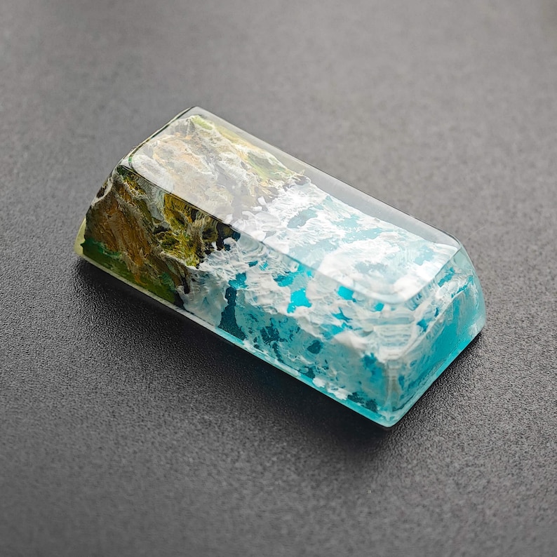 May include: A rectangular, clear resin keycap with a blue and white wave design and a green and brown island design.