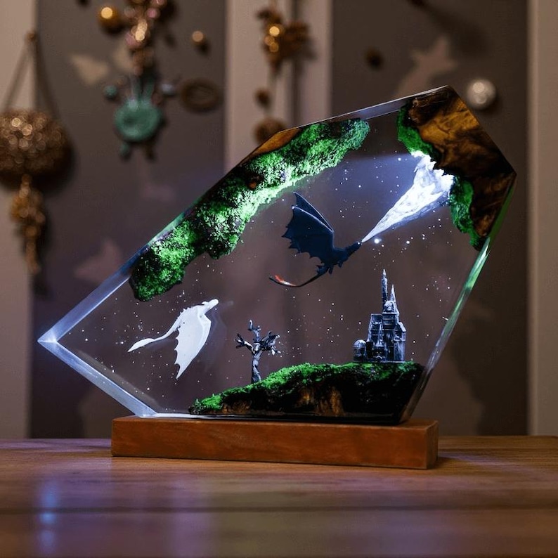可能包含：一件奇幻主题的树脂艺术品，描绘了一条黑龙向一条白龙喷火的场景。画面中包含一座城堡、一棵树和长满青苔的绿色地面，所有元素都以繁星点点的背景衬托。艺术品安装在木质底座上。