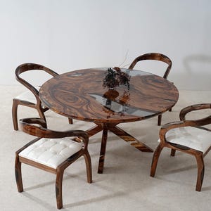 Puede incluir: Una mesa de comedor redonda de madera con un patrón de vetas oscuras y arremolinadas y una incrustación de resina epoxi transparente. Cuatro sillas a juego con marcos de madera curvados y cojines de asiento blancos y acolchados rodean la mesa. Un pequeño jarrón con flores secas está sobre la mesa.