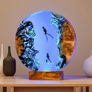 Könnte beinhalten: Ein rundes, beleuchtetes Kunstwerk aus Harz mit einer blauen Unterwasserszene mit zwei Tauchern. Das Kunstwerk ist von Holz und Harz umrahmt und steht auf einem Holzsockel. Der Hintergrund ist ein Blauton.