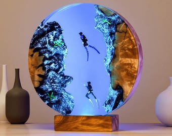 Könnte beinhalten: Ein rundes, beleuchtetes Kunstwerk aus Harz mit einer blauen Unterwasserszene mit zwei Tauchern. Das Kunstwerk ist von Holz und Harz umrahmt und steht auf einem Holzsockel. Der Hintergrund ist ein Blauton.