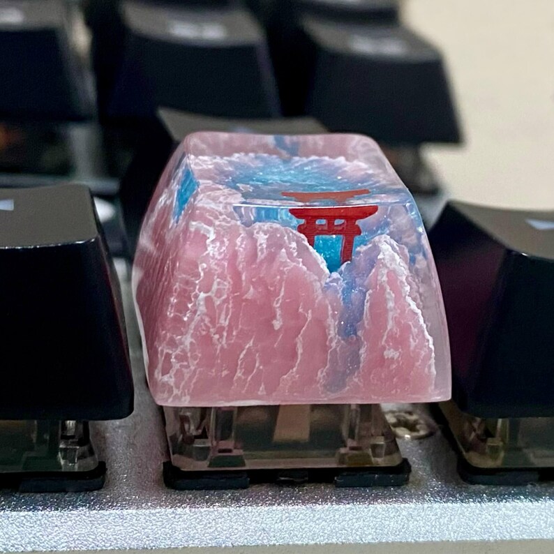 May include: A custom keyboard keycap featuring a pink mountain range design with a red torii gate and blue sky. The keycap is made of resin and sits among other black keycaps on a silver keyboard.