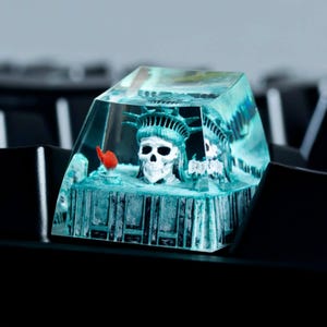 Peut inclure: Un capuchon de touche en résine transparente avec une sculpture miniature de la Statue de la Liberté à l'intérieur. La statue est peinte en vert et blanc, et la base du capuchon de touche est de couleur vert foncé.