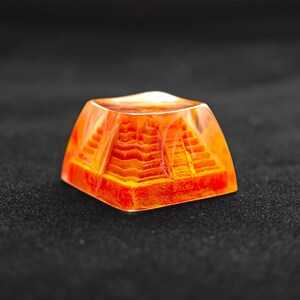 May include: A square, orange-colored pyramid-shaped object encased in clear resin. The pyramid has detailed steps and is illuminated from within. The object is set against a dark background.