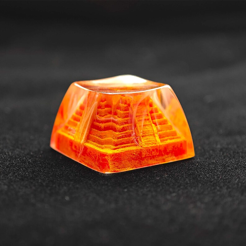 May include: A square, orange-colored pyramid-shaped object encased in clear resin. The pyramid has detailed steps and is illuminated from within. The object is set against a dark background.