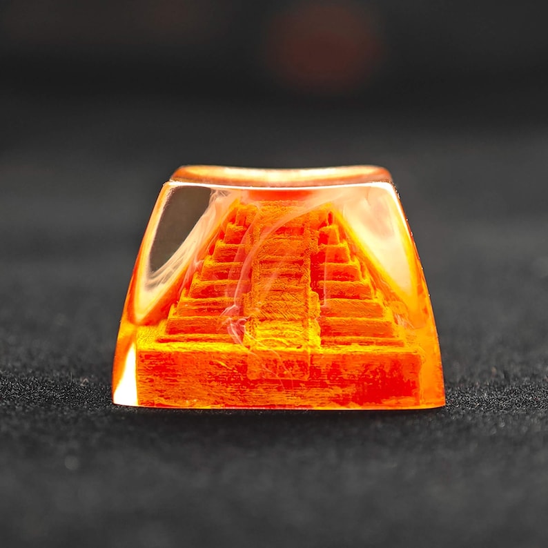 May include: A custom keycap featuring a detailed orange pyramid design encased in clear resin. The pyramid has multiple tiers and steps, creating a 3D effect. The keycap is square-shaped and sits on a dark surface.