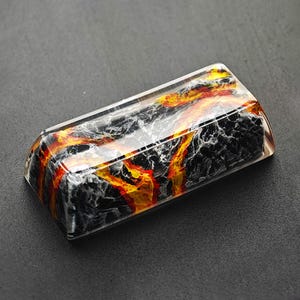 May include: A black and white resin keycap with a red and yellow lava flow design.