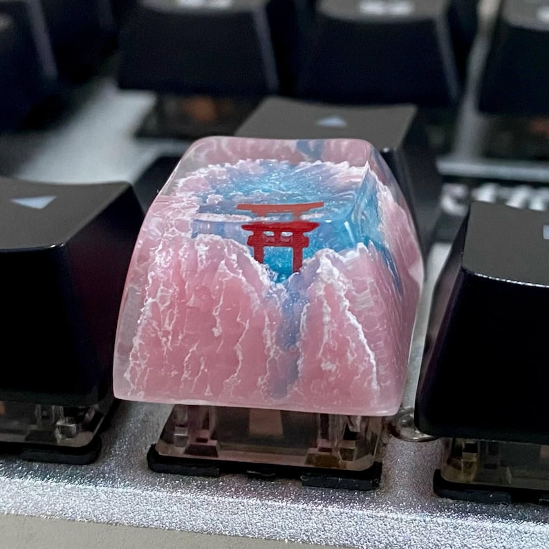 May include: A custom keycap featuring a Japanese torii gate design. The keycap has a pink mountain range and blue sky background. The torii gate is red. The keycap is on a mechanical keyboard.