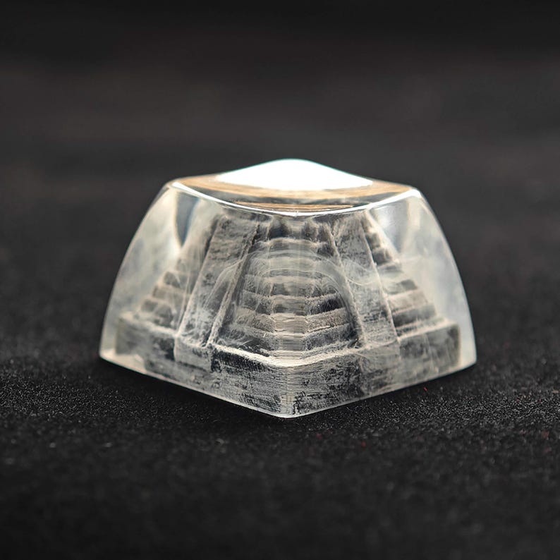 May include: A clear, resin pyramid with a detailed, gray-toned pyramid structure inside. The pyramid is encased in a clear, rounded block, with a white top. The object is displayed on a dark surface.