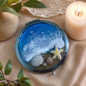 May include: Circular, silver compact mirror with a beach scene design. The mirror features a blue and white wave design, seashells, a yellow starfish, and a pearl. The compact is a decorative item.