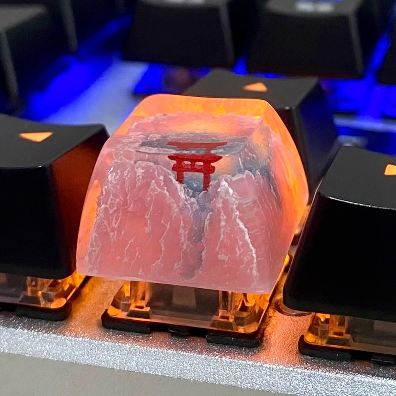 May include: Close-up of a custom keyboard keycap featuring a red Torii gate design set against a mountain landscape. The keycap is translucent with a pink and orange gradient, and is surrounded by black keycaps.