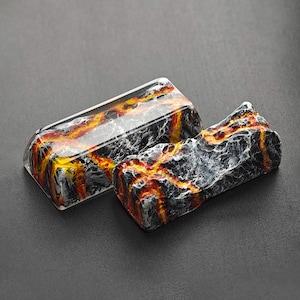 May include: Two keycaps with a lava rock design. One keycap is clear with a black and orange lava rock design inside. The other keycap is opaque and has a black, orange, and white lava rock design.