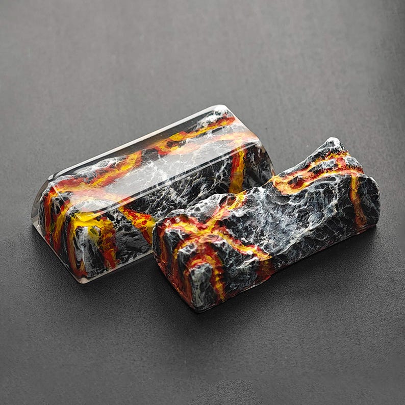 May include: Two keycaps with a lava rock design. One keycap is clear with a black and orange lava rock design inside. The other keycap is opaque and has a black, orange, and white lava rock design.