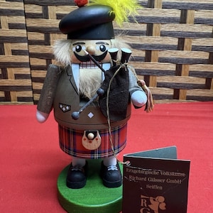 May include: A wooden nutcracker figurine dressed in a kilt and tam o' shanter with a yellow feather, holding bagpipes. It stands on a green base next to a black tag labeled Erzgebirgische Volkskunst Richard Glässer GmbH Seiffen.