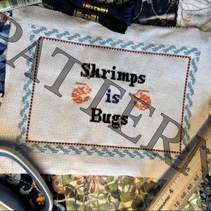 May include: A white cross-stitch embroidery with a blue and red border. The embroidery says "Shrimps is Bugs".