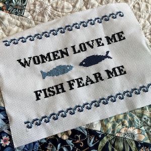 May include: White cross-stitch embroidery with blue and black thread. The text reads "Women Love Me Fish Fear Me" with two fish designs.