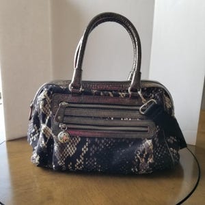 May include: A black and white snakeskin-patterned handbag with silver-toned accents. The bag features a top handle, multiple zippered compartments, and a detachable black shoulder strap. A decorative charm hangs from the front.
